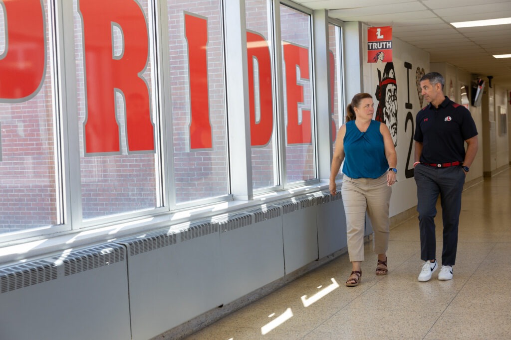 Two educators walk side by side down a school hallway engaged in conversation, as they walk past a window adorned with large letters that spell "pride" for school pride. 