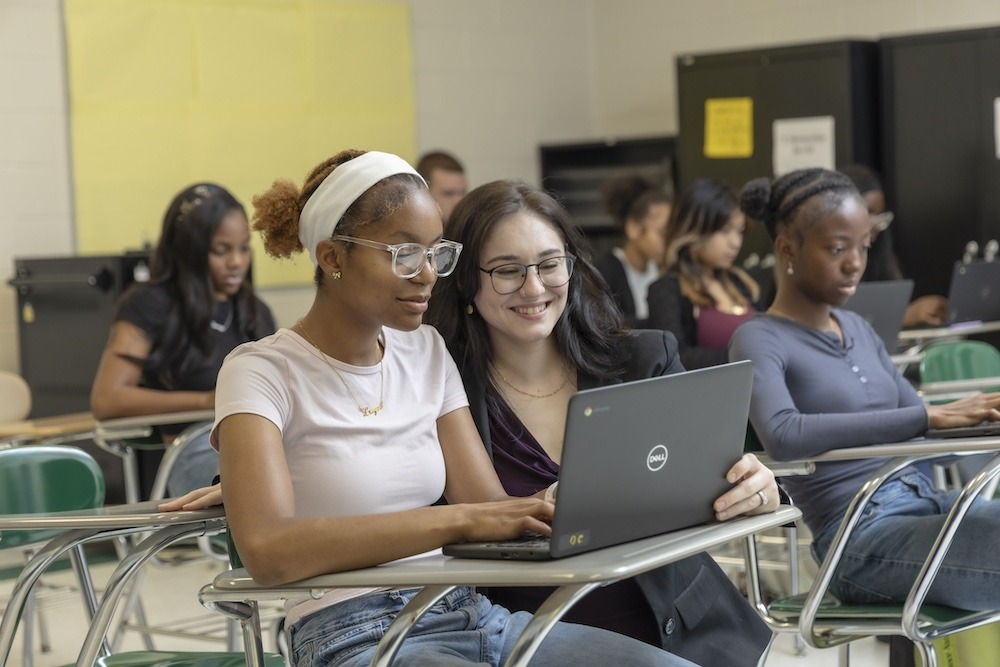 An educator crouches next to a student in her class to review a concept on the student's laptop. 