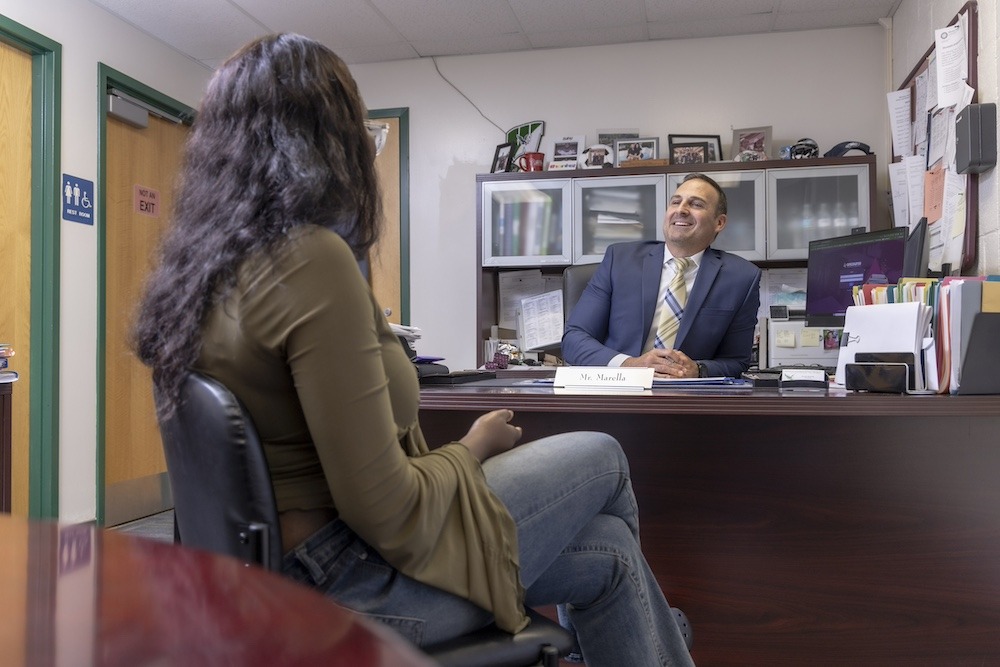 A principal sits at his desk as a student visits his office to talk to him, sitting across from him. 