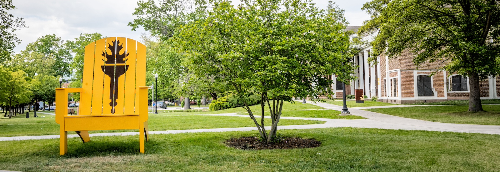 An oversized Rowan University yellow Adirondack chair with a Rowan brown torch painted on it sits on the grass on campus.