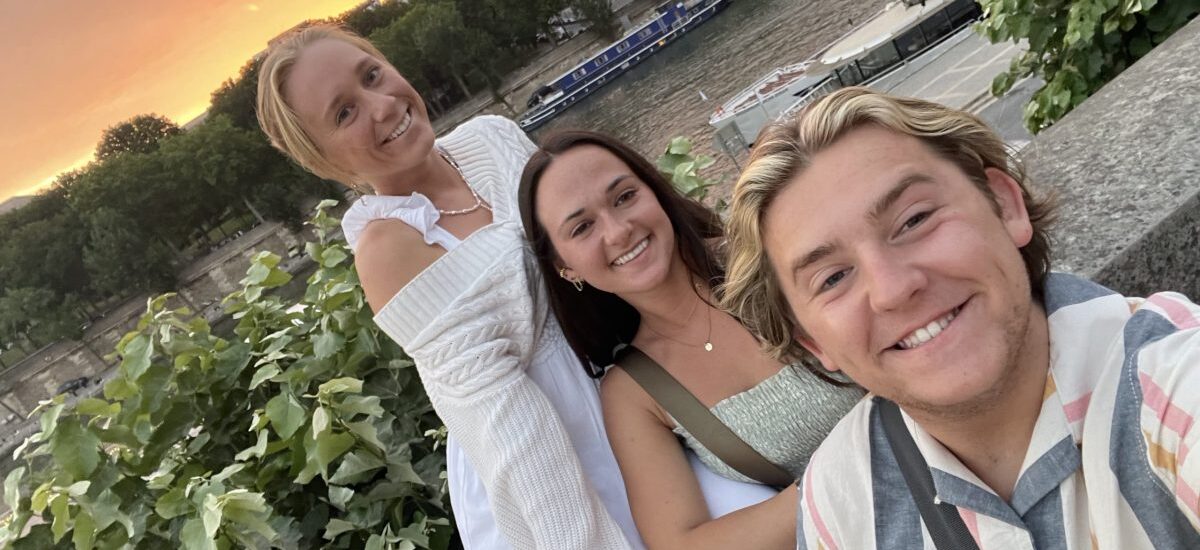Three college students taking a photo together near a river during summer travel.