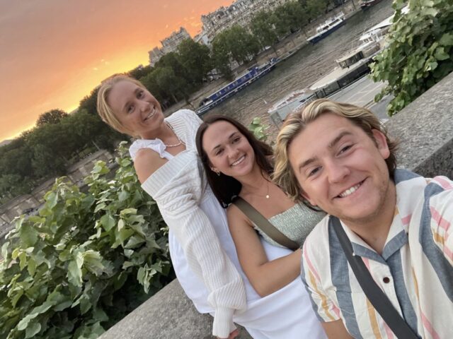 Three college students taking a photo together near a river during summer travel.