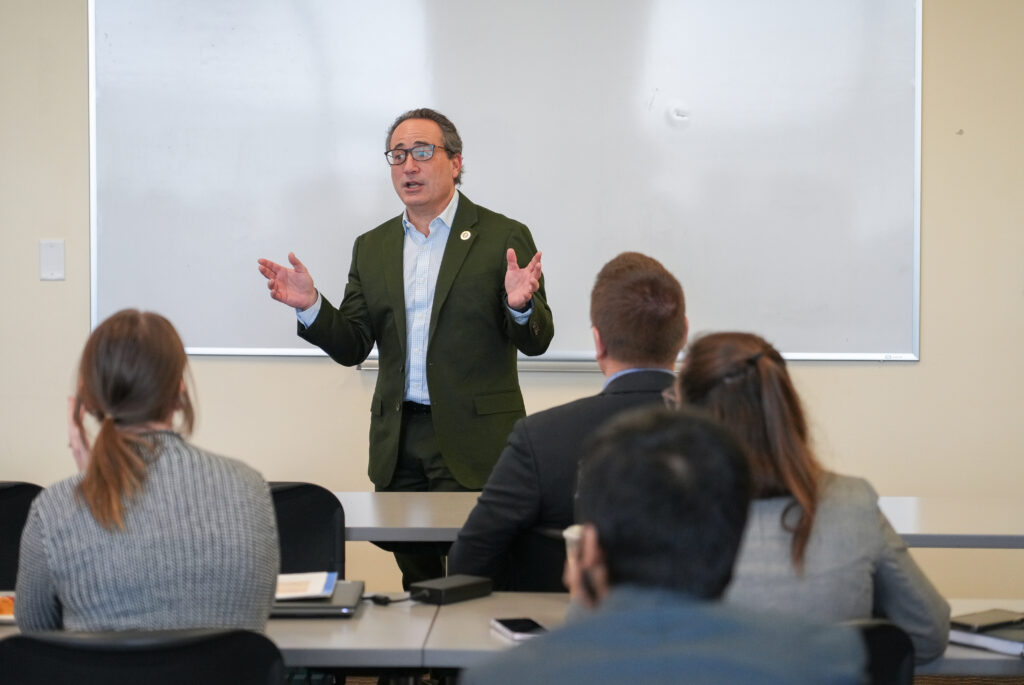 A speaker stands at the front of a classroom and presents to a group of seated students and professionals during a leadership-focused discussion.