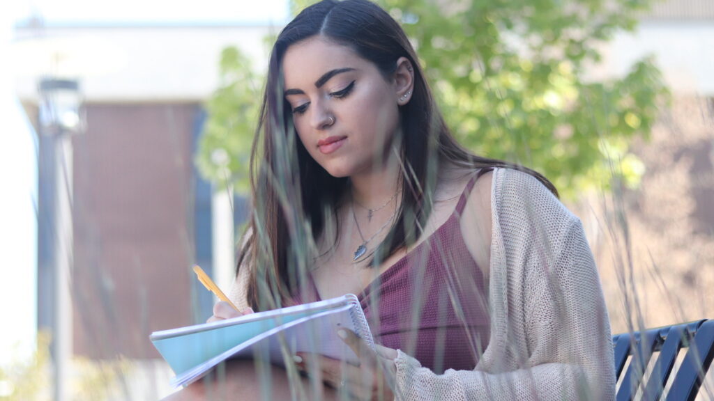 A student sits outdoors and writes in a notebook, representing continuous learning, reflection and personal growth.