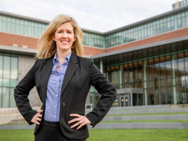 Woman in a dark blazer and blue shirt stands with her hands on her hips outside a Rowan University building.