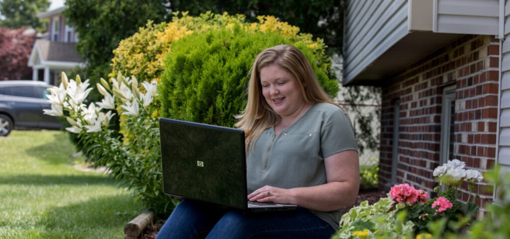 Female MBA candidate sitting outside near a garden and brick house while working on a laptop.
