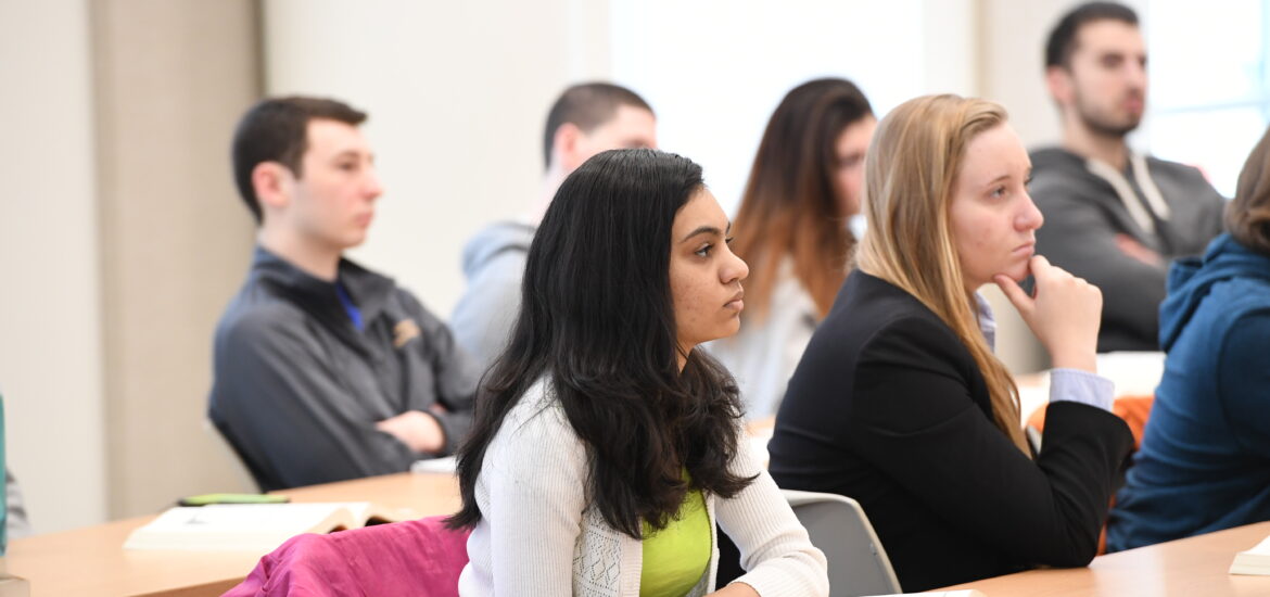 Students sit at desks in a classroom and listen during a business class discussion, representing learning, focus and professional skill development.