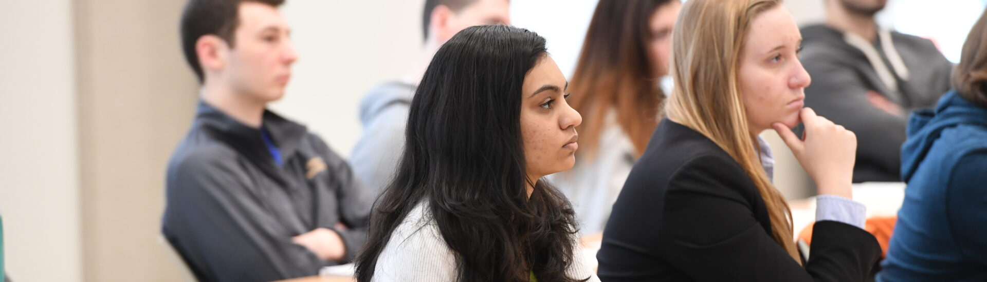 Students sit at desks in a classroom and listen during a business class discussion, representing learning, focus and professional skill development.