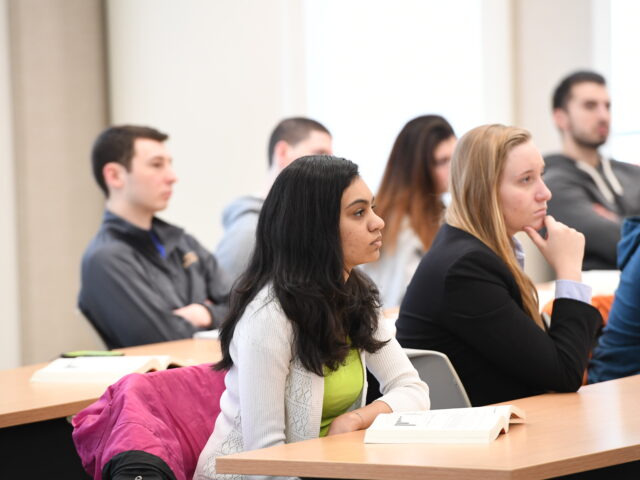 Students sit at desks in a classroom and listen during a business class discussion, representing learning, focus and professional skill development.