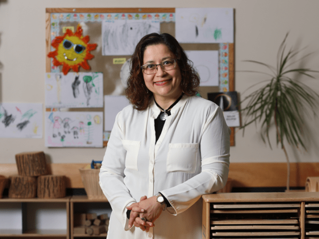 Teacher standing in a bright classroom in front of student artwork, learning materials and wooden classroom shelves.