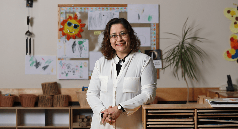 Teacher standing in a bright classroom in front of student artwork, learning materials and wooden classroom shelves.