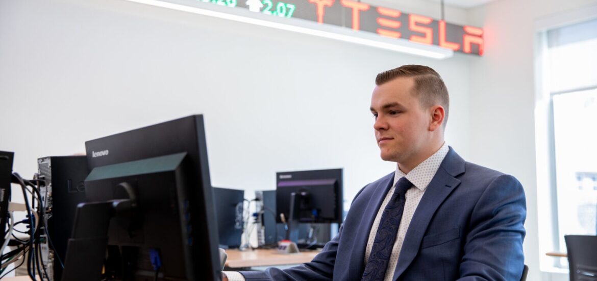 Business administration student in a suit working at a computer in a finance lab with stock ticker displays overhead.