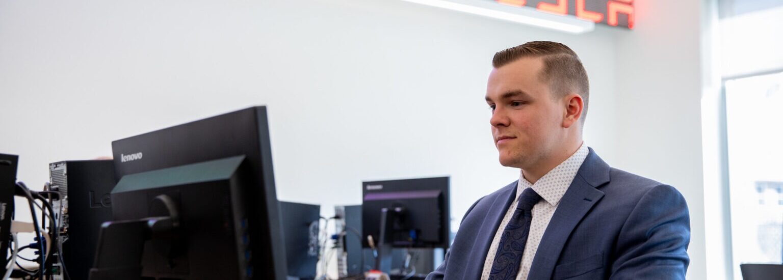 Business administration student in a suit working at a computer in a finance lab with stock ticker displays overhead.