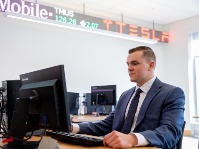 Business administration student in a suit working at a computer in a finance lab with stock ticker displays overhead.