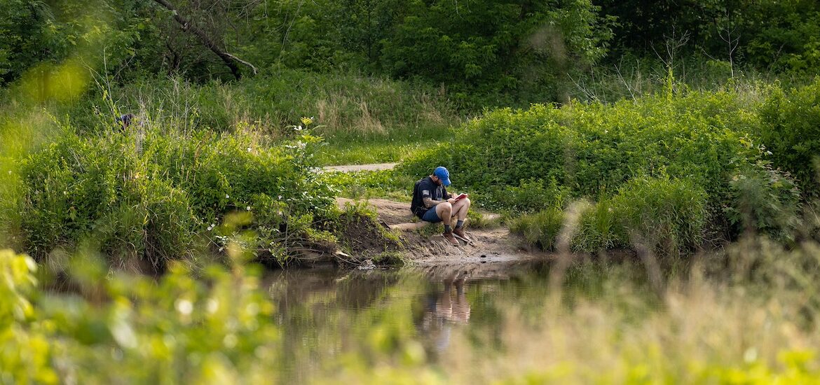 A Rowan University students sits on a riverbank in the Pine Barrens, surrounded by trees and water.