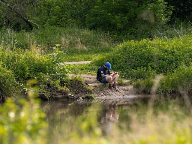 A Rowan University students sits on a riverbank in the Pine Barrens, surrounded by trees and water.