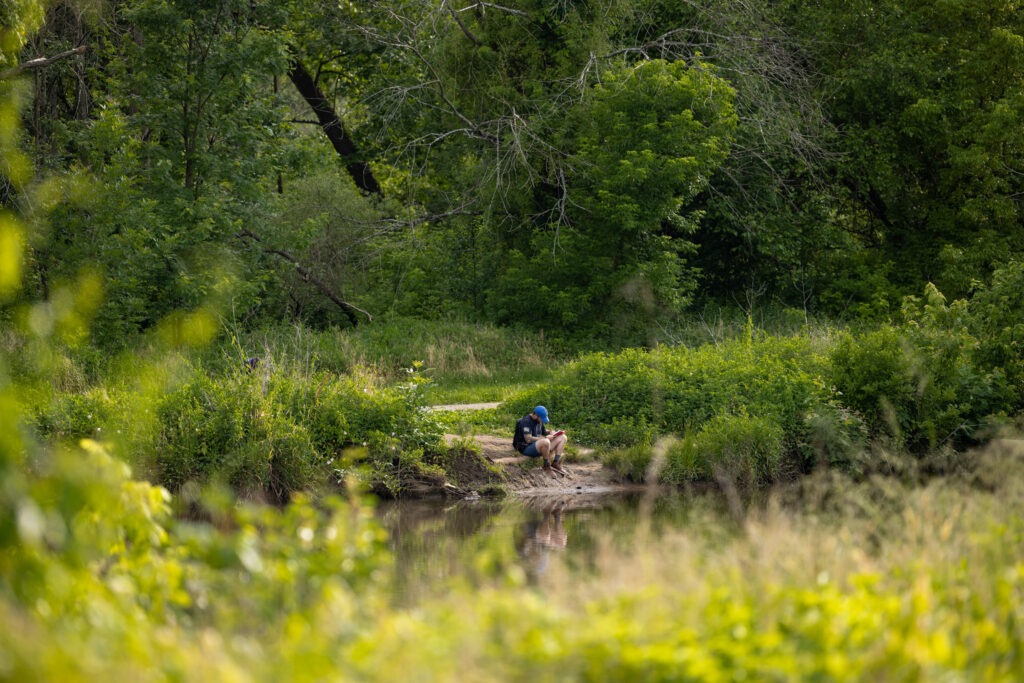 A Rowan University student sits on an embankment on the other side of a body of water, his knees tucked up and he is concentrating on something he is writing. We see him surrounded by greenery and looking small in nature.
