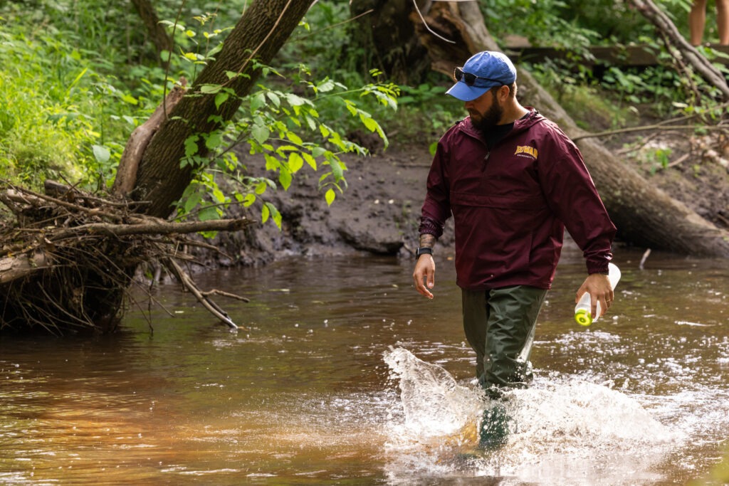 A Rowan University student walks through a shallow part of water in the Pine Barrens, creating splashes around him.