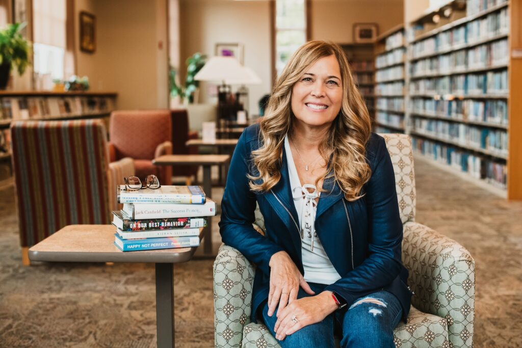 Rowan University online psychology graduate Cindy C. sits in a library for a posed portrait, sitting in an armchair next to an end table with a stack of psychology books and a pair of glasses. 