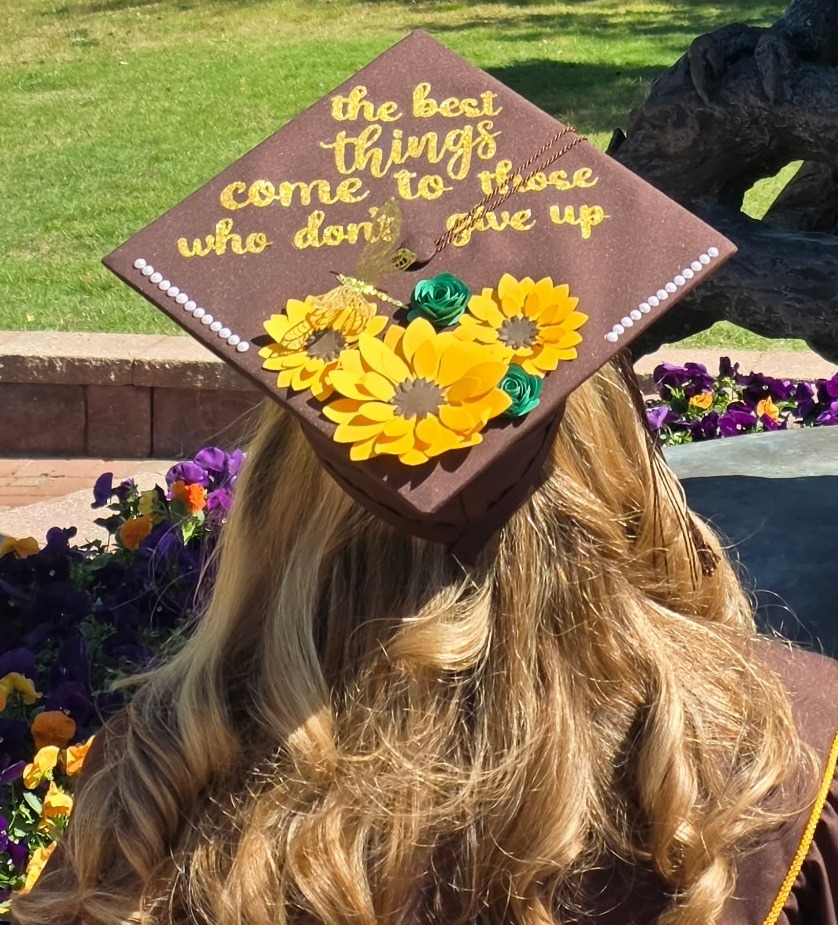 Rowan University online psychology graduate Cindy C. as seen from behind. This is a close up of her decorated graduated cap, which says "The best things come to those who don't give up" and is adorned with three black eyed Susan and two green rose blooms. 