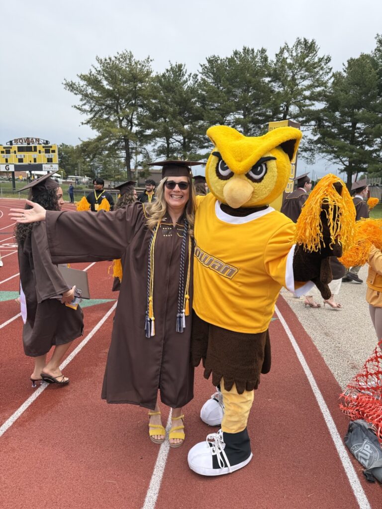 Rowan University online psychology graduate Cindy C. beams with arms outstretched, mirroring the outstretched arms of the Rowan University mascot, Whoo RU, who is by her side, after her graduation ceremony. 