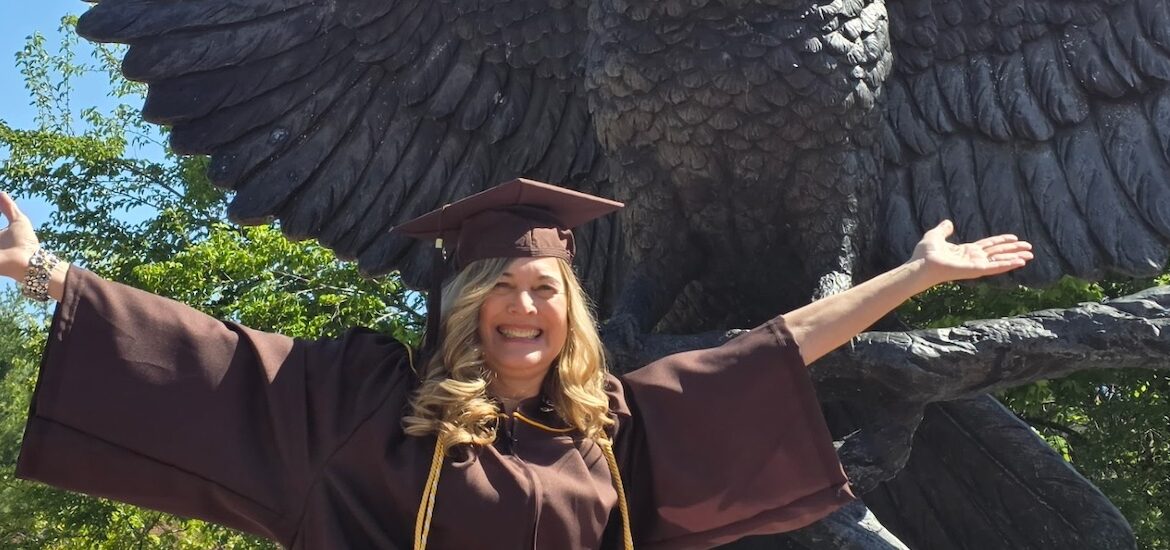 Rowan University online psychology graduate Cindy C. sits in front of the campus statue of an owl with its wings outstretched, matching the owl's pose wit her own outstretched arms and a big smile wearing her graduation cap and gown.