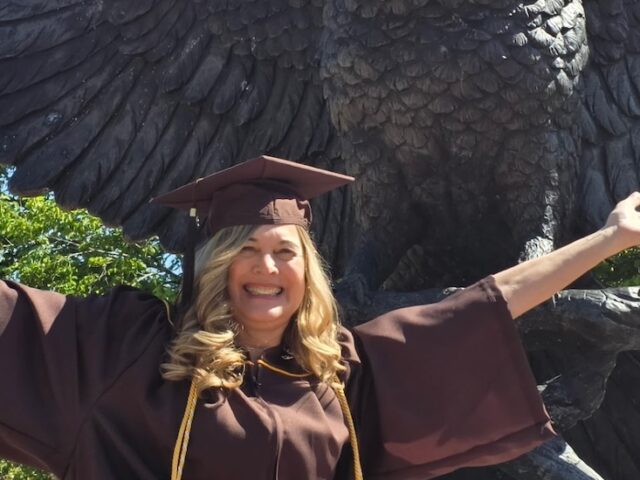 Rowan University online psychology graduate Cindy C. sits in front of the campus statue of an owl with its wings outstretched, matching the owl's pose wit her own outstretched arms and a big smile wearing her graduation cap and gown.