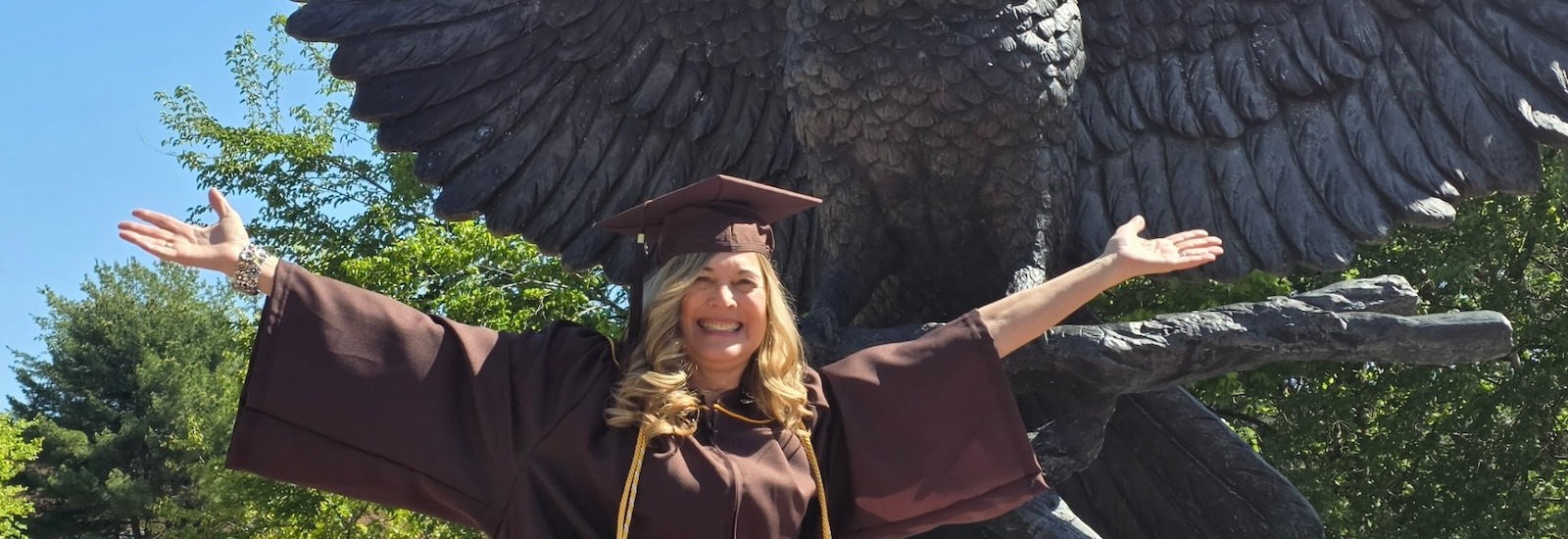Rowan University online psychology graduate Cindy C. sits in front of the campus statue of an owl with its wings outstretched, matching the owl's pose wit her own outstretched arms and a big smile wearing her graduation cap and gown.