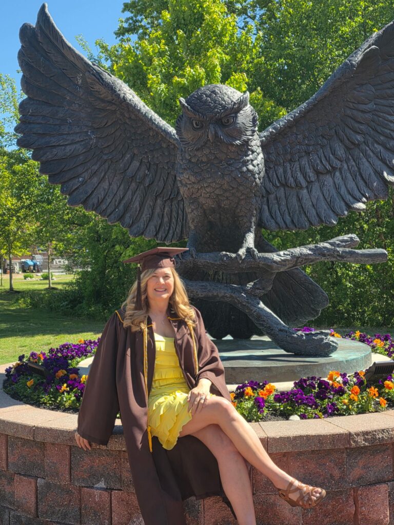 Rowan University online psychology graduate Cindy C. sits on the concrete ring around the statue of an owl with outstretched wings. She is wearing her brown graduation gown, draped open showing her yellow dress and highlighting the yellow of her graduation cords. 