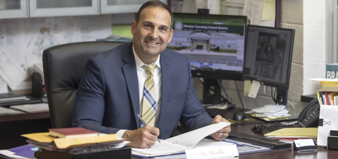 A school administrator in a navy suit sits at his office desk holding paperwork, with school district webpages displayed on two computer monitors behind him.