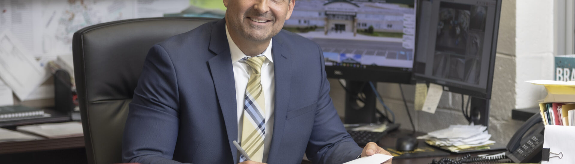 A school administrator in a navy suit sits at his office desk holding paperwork, with school district webpages displayed on two computer monitors behind him.