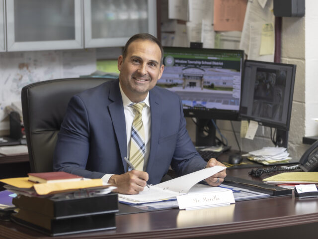 A school administrator in a navy suit sits at his office desk holding paperwork, with school district webpages displayed on two computer monitors behind him.