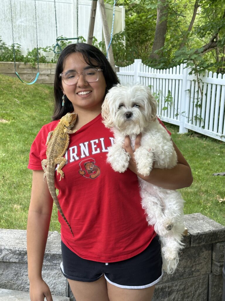 Rowan University biological sciences graduate Kayla B. stands in a backyard wearing a Cornell t-shirt, holding a white, fluffy dog in her left arm and an iguana on her right shoulder.