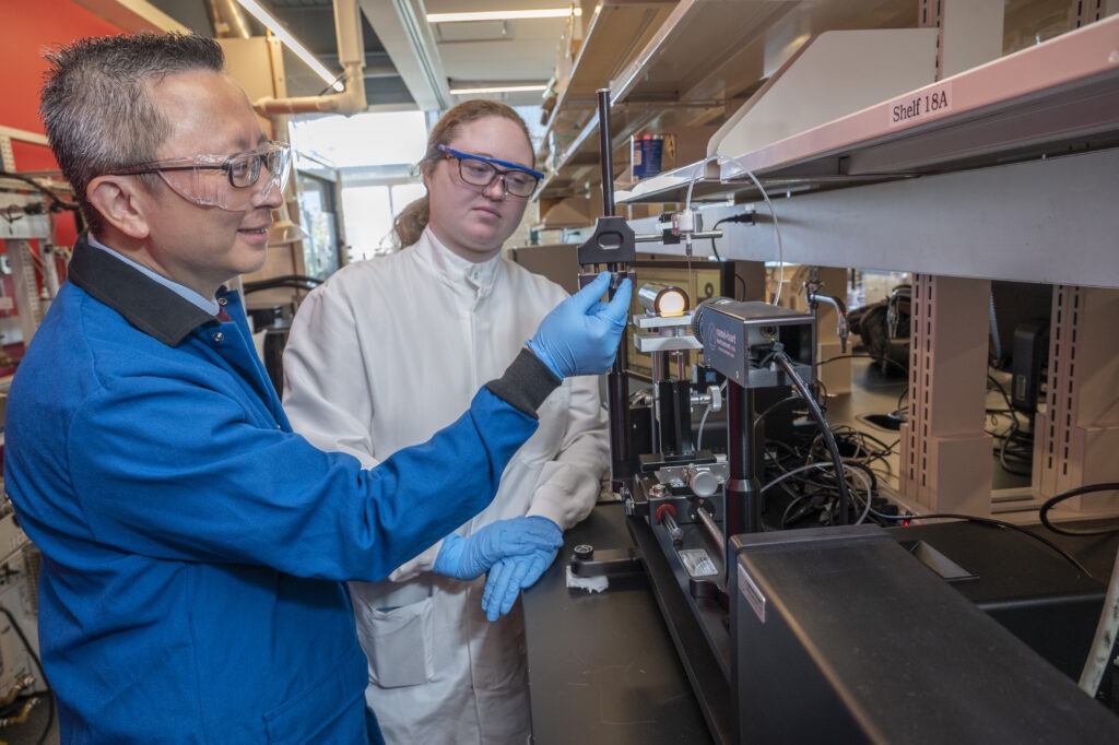 Engineering faculty member wearing safety glasses demonstrates lab equipment to a student in a research laboratory.