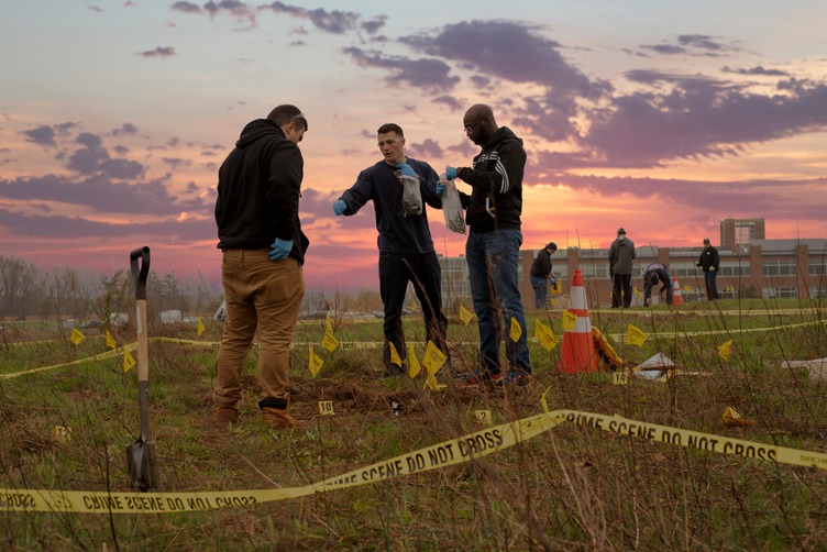 Three emergency response professionals wearing gloves examine a marked outdoor scene behind crime-scene tape while other team members work nearby.