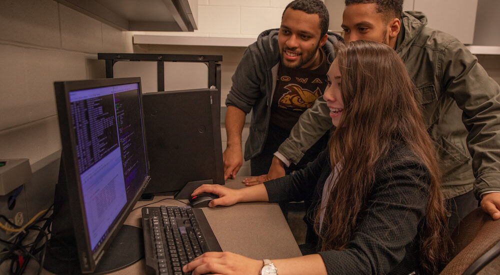 Three students gather around a desktop computer while one student uses the mouse to review lines of code on the screen.