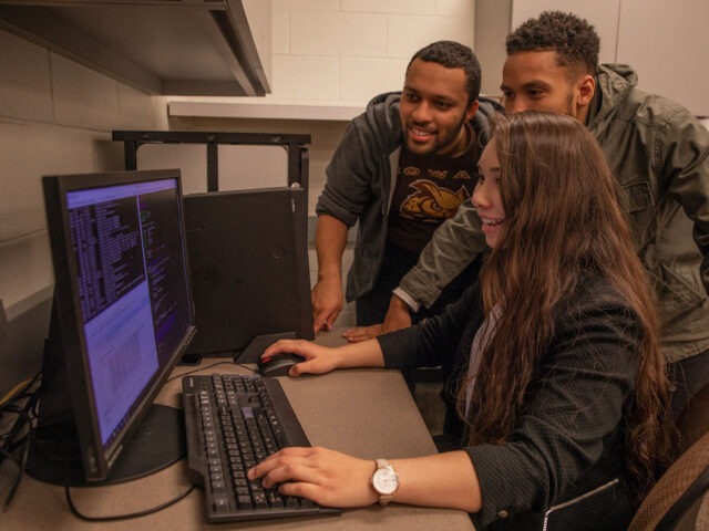 Three students gather around a desktop computer while one student uses the mouse to review lines of code on the screen.