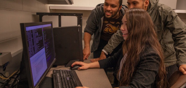 Three students gather around a desktop computer while one student uses the mouse to review lines of code on the screen.