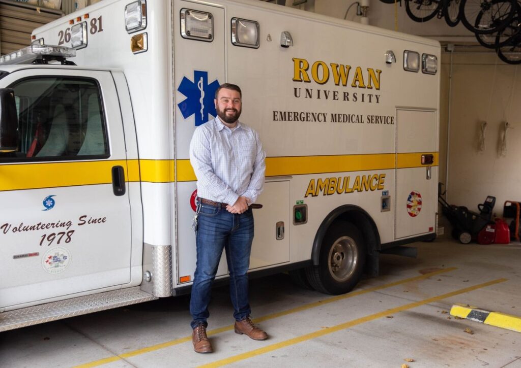 A man stands beside a Rowan University Emergency Medical Service ambulance inside a vehicle garage.