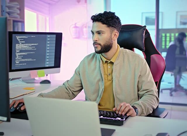 A man works at a desk with a laptop, keyboard and multiple monitors displaying computer code in a modern office.