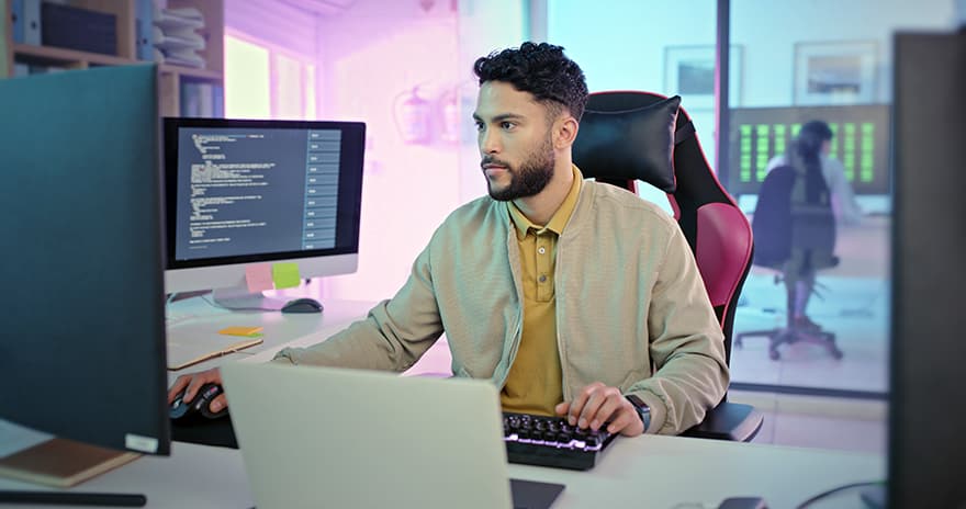 A man works at a desk with a laptop, keyboard and multiple monitors displaying computer code in a modern office.