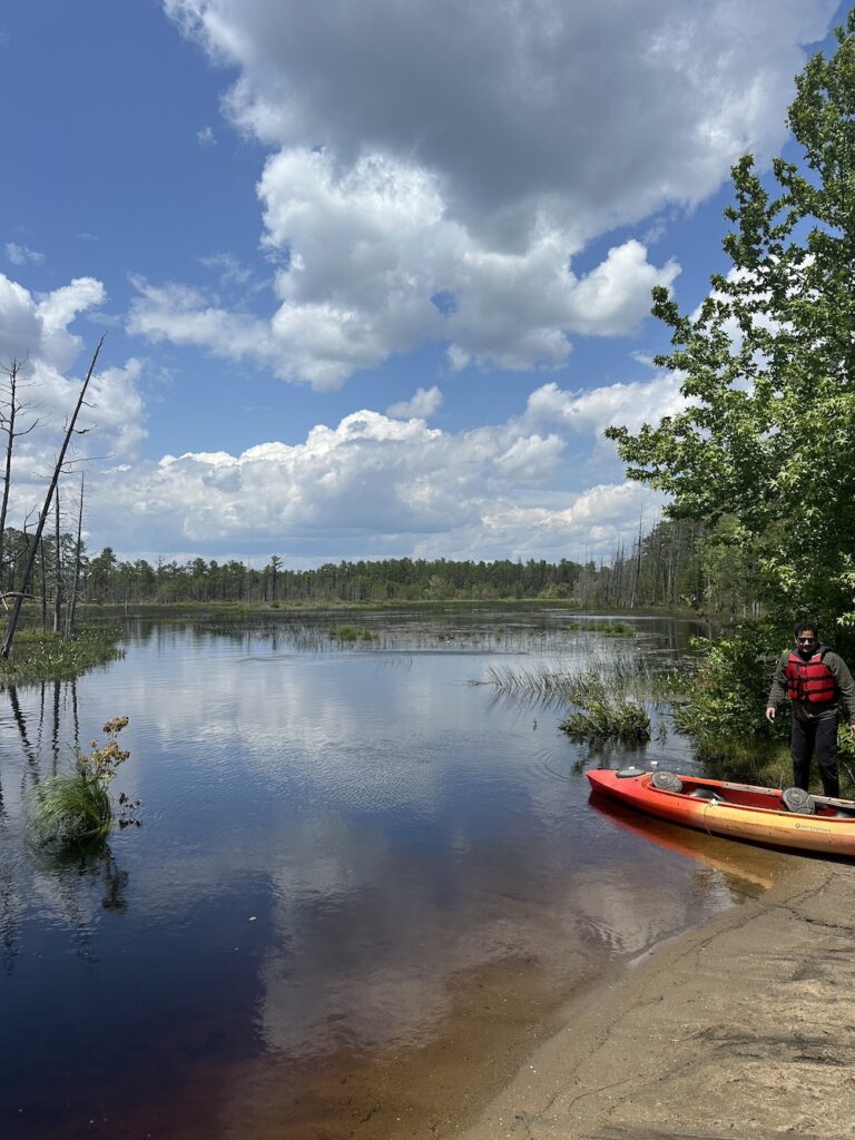 Big, poofy white clouds in a bright blue sky reflect off of the water of a wide, mostly still river in the Pine Barrens. There is a man wearing a life vest pulling his kayak in off the water.