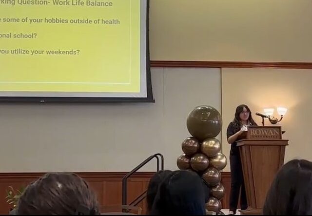Rowan University biological sciences major Kayla B. stands at a Rowan University podium, delivering spoken remarks to a seated crowd.