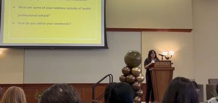 Rowan University biological sciences major Kayla B. stands at a Rowan University podium, delivering spoken remarks to a seated crowd.