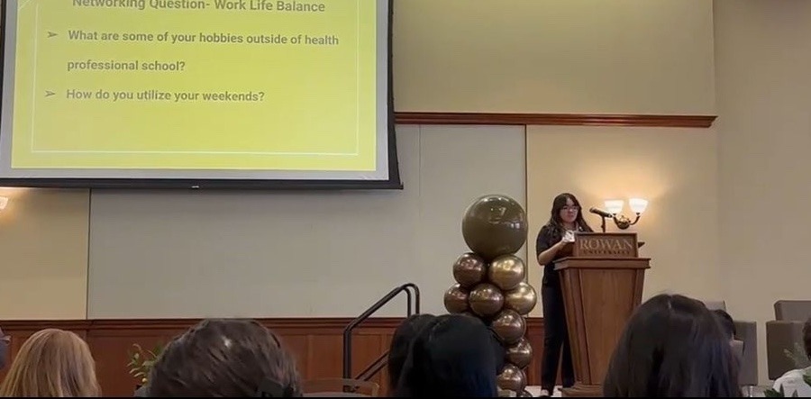 Rowan University biological sciences major Kayla B. stands at a Rowan University podium, delivering spoken remarks to a seated crowd.