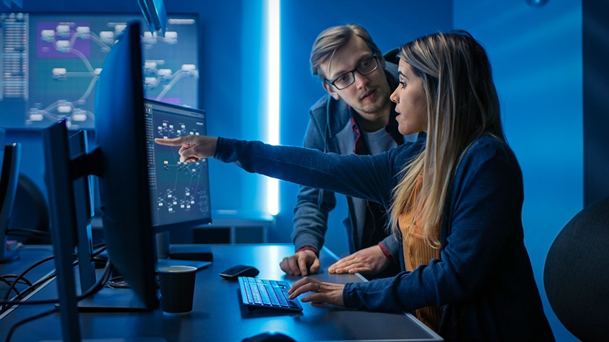A woman points to data visualizations on a computer monitor while a man looks on in a blue-lit technology lab.