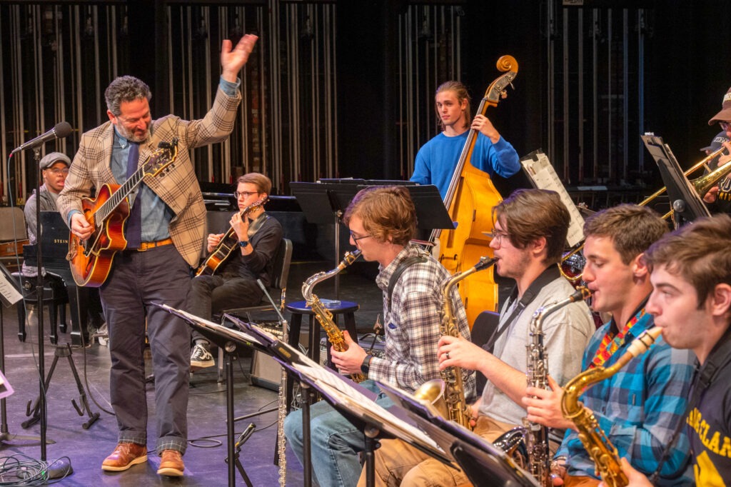 A jazz ensemble performs onstage as a guitarist leads musicians playing saxophones and upright bass.