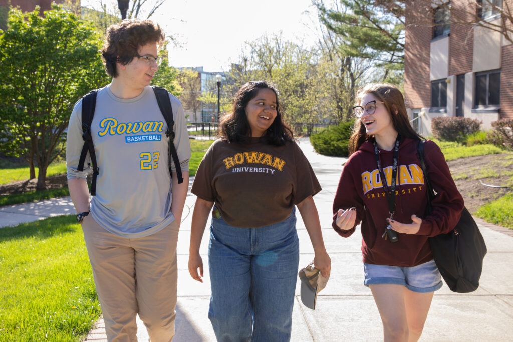 Three Rowan University students wearing Rowan apparel walk together and talk on a campus sidewalk.