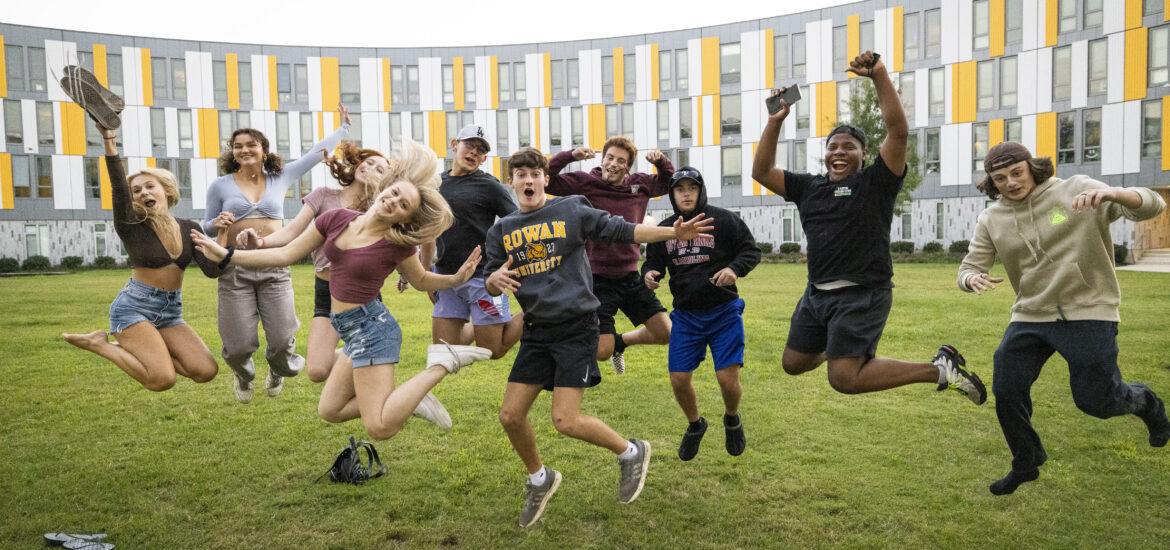 A group of Rowan University students jump and pose together on a grassy lawn outside a campus residence hall.