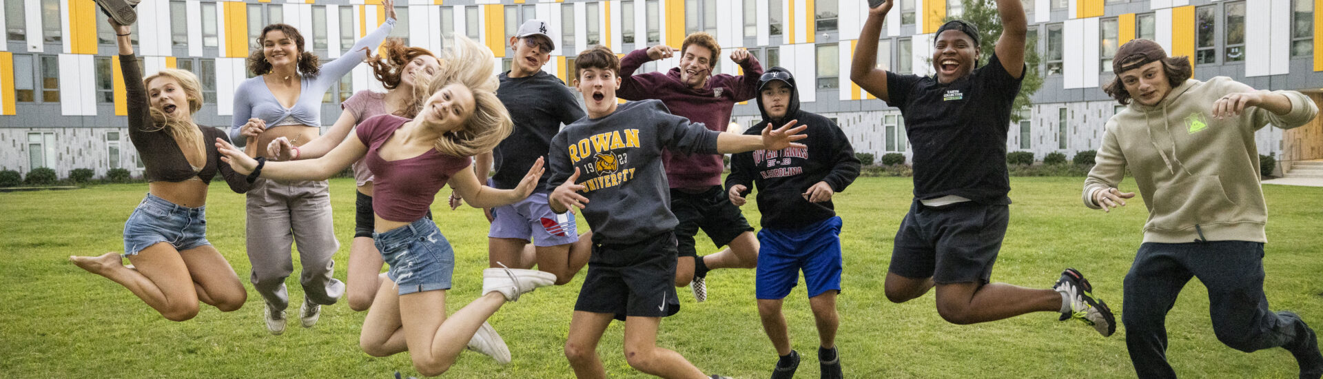 A group of Rowan University students jump and pose together on a grassy lawn outside a campus residence hall.
